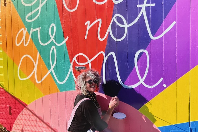 Photo of Sarah Johnson painting a colorful "you are not alone" mural.