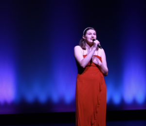 Photo of a young woman wearing a red dress and singing into a microphone.