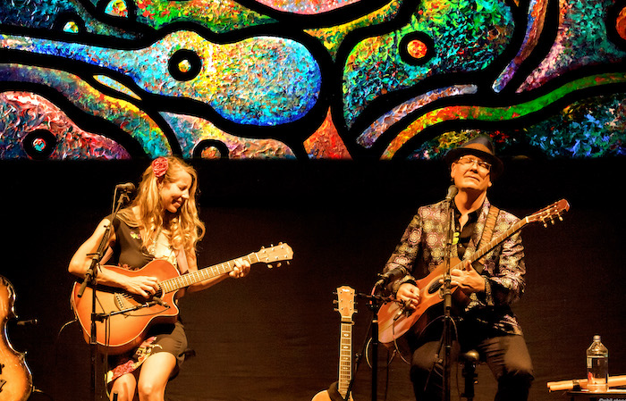 Photo of Patchouli & Terra Guitarra performing on guitars with bright-colored guitar imagery behind them.