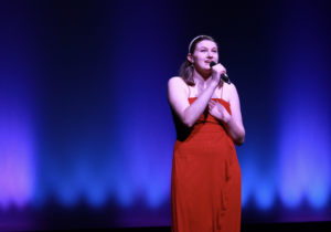 Photo of a young woman wearing a red dress and singing into a microphone.