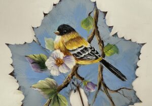 A painting by Barb Halvorson done on a leaf, showing a goldfinch and an apple blossom.