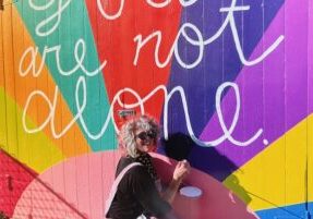 Photo of Sarah Johnson painting a colorful "you are not alone" mural.