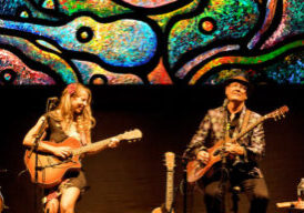 Photo of Patchouli & Terra Guitarra performing on guitars with bright-colored guitar imagery behind them.